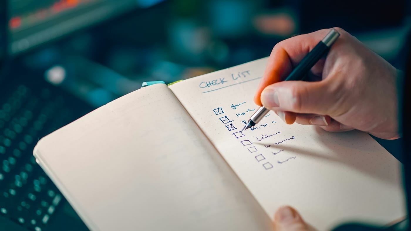 Mão preenchendo um checklist de processo de vendas em um caderno, com etapas de atendimento e qualificação de leads