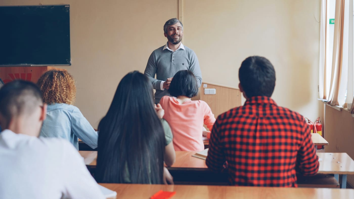Professor dando aula para uma turma de escola de idiomas, com os alunos sentados de costas para a câmera