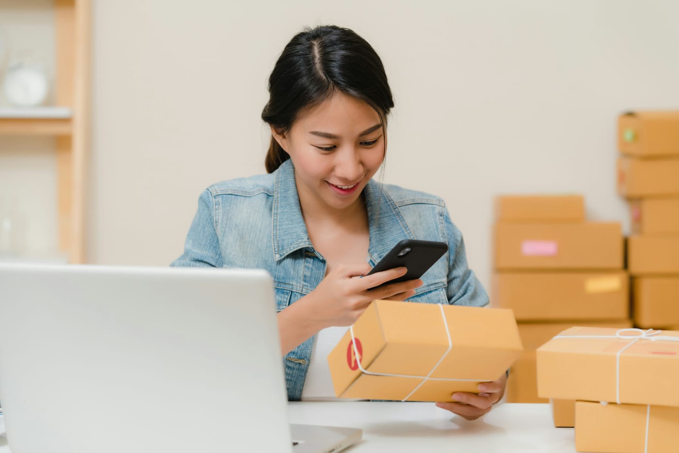 Empreendedora sorrindo ao conferir um pedido no celular, cercada de caixas de encomenda prontas para envio